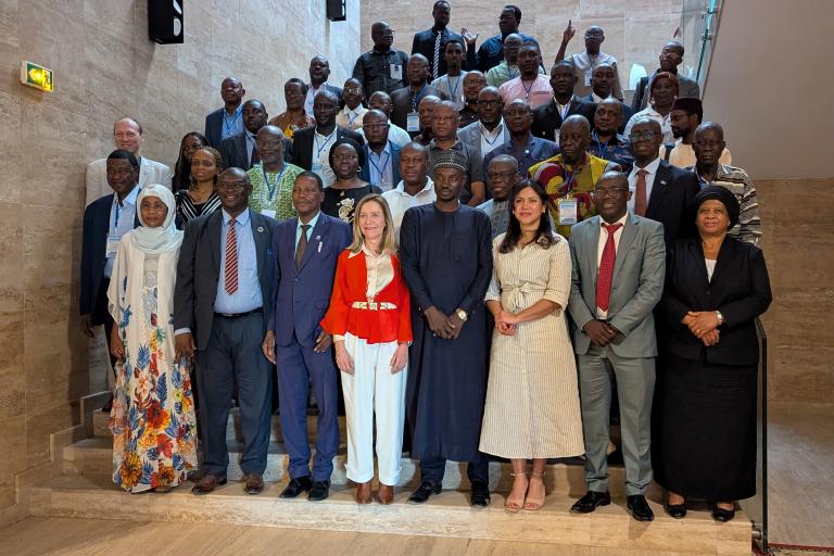 A large group of people, dressed in formal and traditional attire, pose for a group photo on a staircase in an indoor setting.