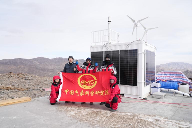 Four people in red jackets hold a red flag with Chinese text and "AMS" logo in front of a solar- and wind-powered research station on a rocky mountain.