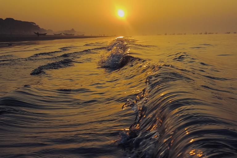 Golden sunlight reflects off gentle ocean waves near the shore at sunrise. Boats and silhouettes are visible in the distance under an orange sky.