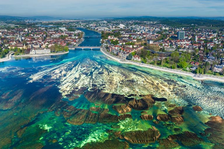 Aerial view of a city with a river flowing into a large lake, showing clear turquoise water, rocky formations, and surrounding urban and green areas.