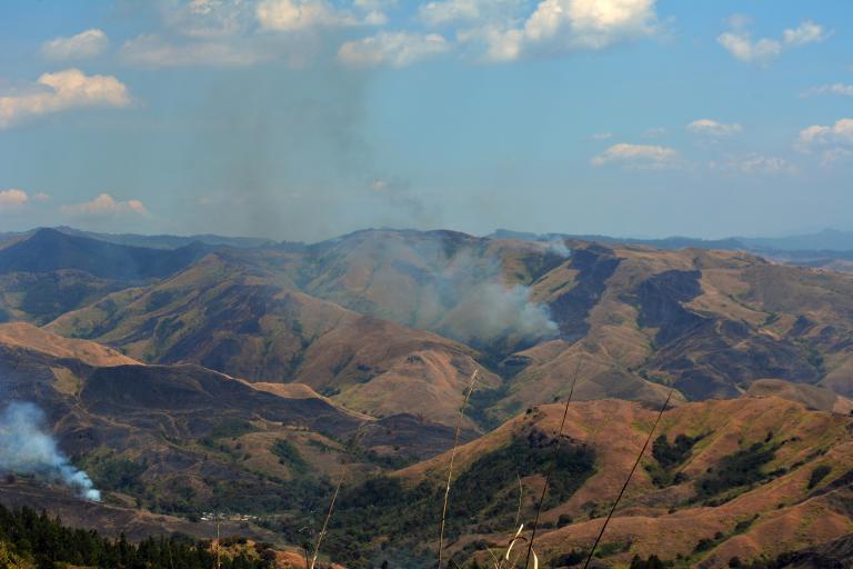 A mountainous landscape with brown, dry hills and scattered smoke rising from multiple areas, under a partly cloudy sky.