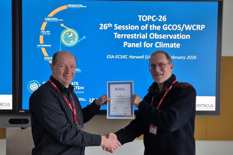 Two men shaking hands, one holding a certificate, in front of a screen displaying information about the 26th Session of the GCOS/WCRP Terrestrial Observation Panel for Climate.