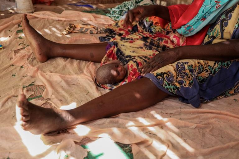 A newborn baby lies on a cloth between an adult woman's legs, with both resting on a patterned fabric floor.