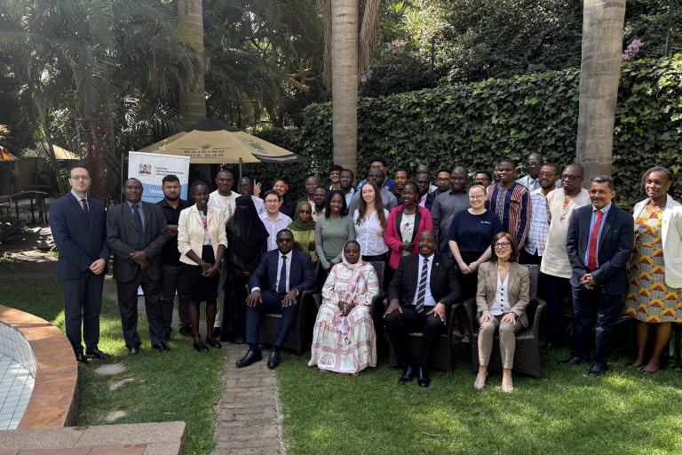 A diverse group of people pose for a formal group photo outdoors, with some seated and others standing, surrounded by greenery and palm trees.