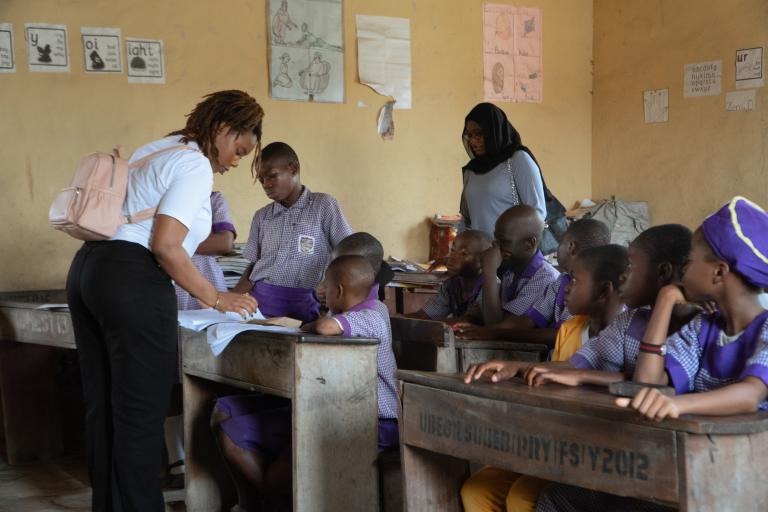 A teacher assists students in a classroom as another adult observes; students are seated at wooden desks wearing purple uniforms. Educational posters are on the walls.