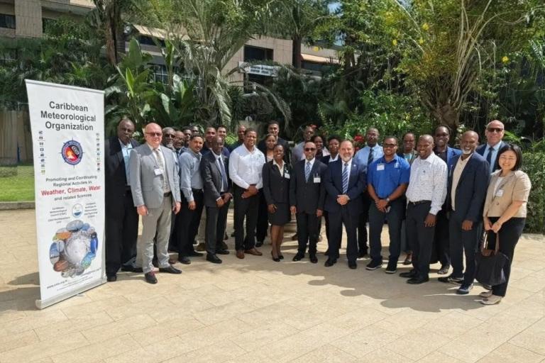 A group of people in formal attire stands outdoors next to a banner for the Caribbean Meteorological Organization. Trees and buildings are visible in the background.