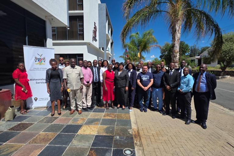 A large group of people posing for a group photo outdoors in front of a building with a banner reading "Meteorological Services.