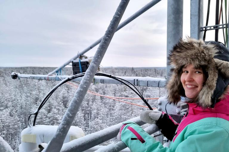 A person in a fur-lined hooded winter coat stands on a metal structure overlooking a snowy forest, smiling at the camera.