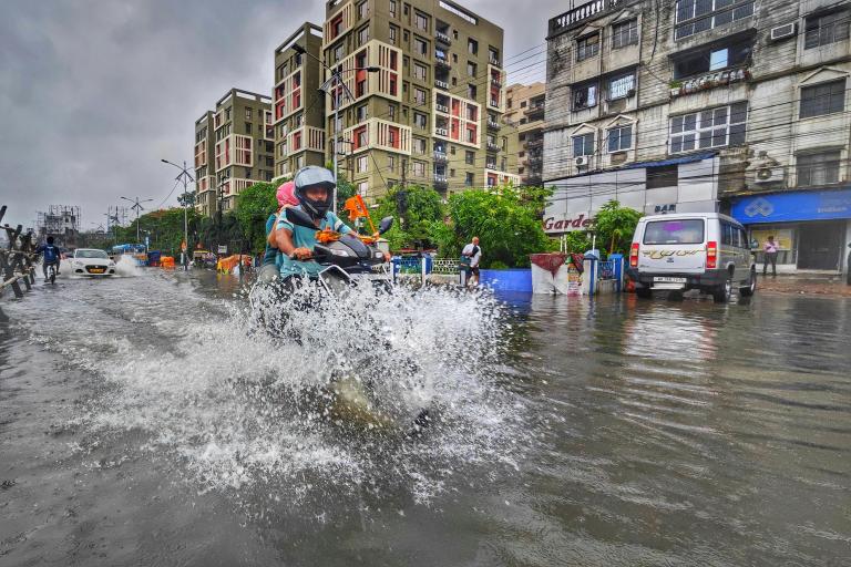 A person rides a motorcycle through a flooded city street, splashing water, with buildings and vehicles in the background on a cloudy day.