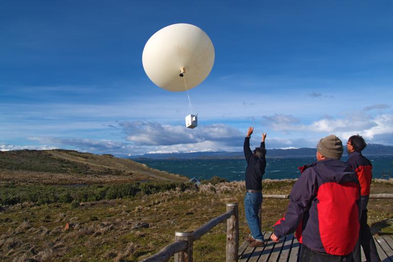 Three people on a wooden platform release a large weather balloon with attached instruments into a clear blue sky over a hilly, grassy landscape.