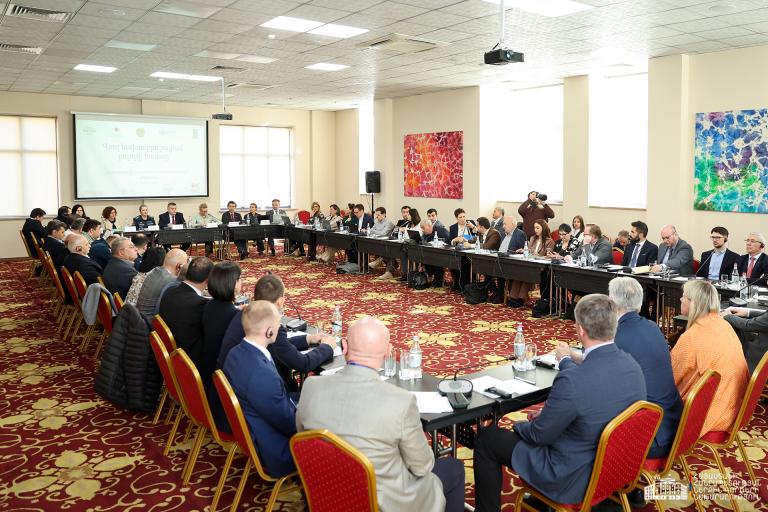 A large group of people sit around a U-shaped conference table in a meeting room, with a presentation slide displayed at the front.