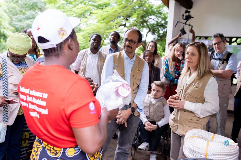 A group of people, some wearing vests, listen attentively to a person in a red shirt holding supplies during an outdoor gathering.