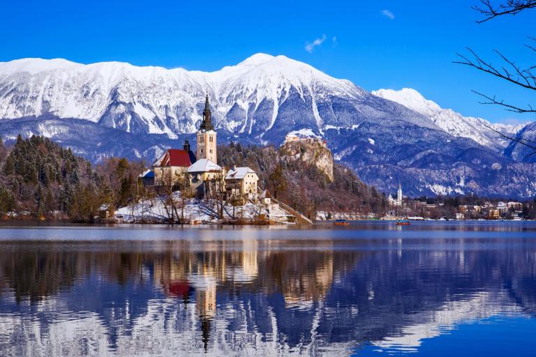 A church and castle stand on Lake Bled’s island, surrounded by snow-capped mountains and reflected in the calm water under a clear blue sky.