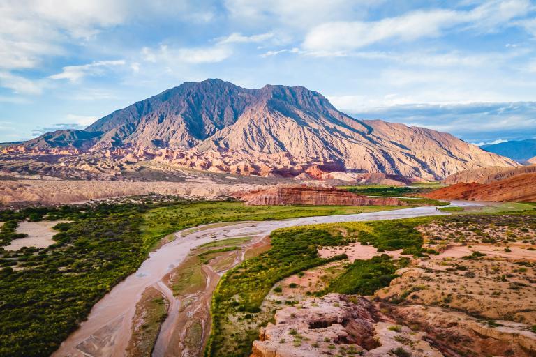 A winding river flows through a green valley with a large, rocky mountain in the background under a partly cloudy sky.