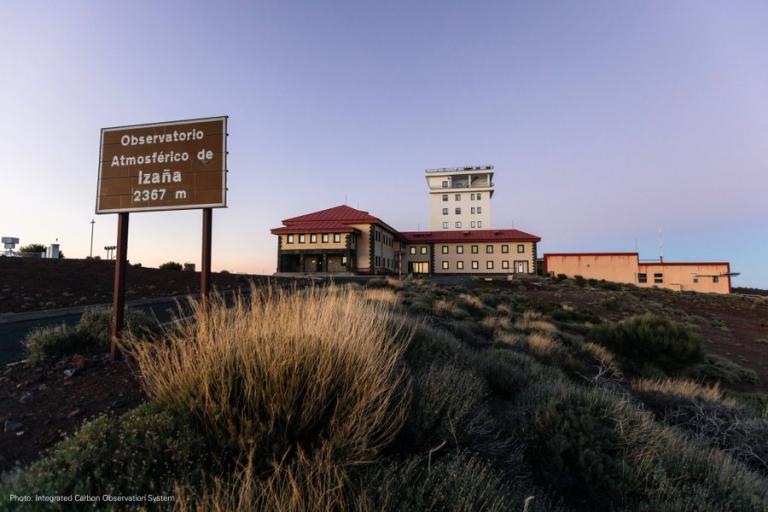 A large building complex sits on a rocky hillside at sunset, with a sign reading "Observatorio Atmosférico de Izaña 2367 m" in the foreground.
