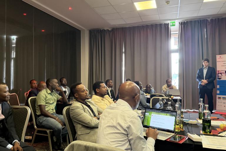 A group of people seated at tables in a conference room listen to a man presenting near a screen and a display banner. Laptops and bottles are on the tables.