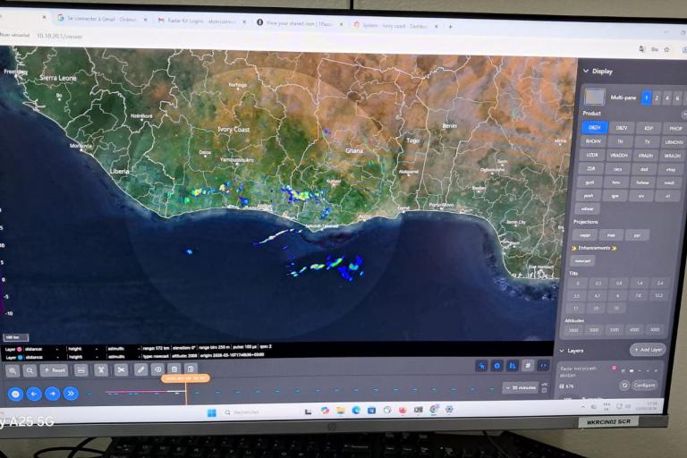 A computer monitor displays a weather radar interface showing precipitation data over a coastal region, with various control panels and map layers visible on the screen.