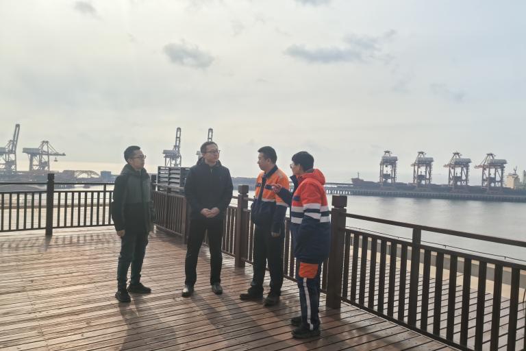 Four people stand on a wooden deck by the water with cranes and industrial structures visible in the background under a cloudy sky.