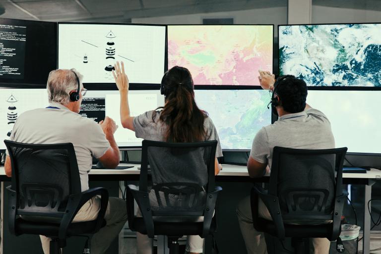 Three people wearing headsets sit at desks, facing large monitors displaying maps, satellite images, and data visualizations in a modern control room.