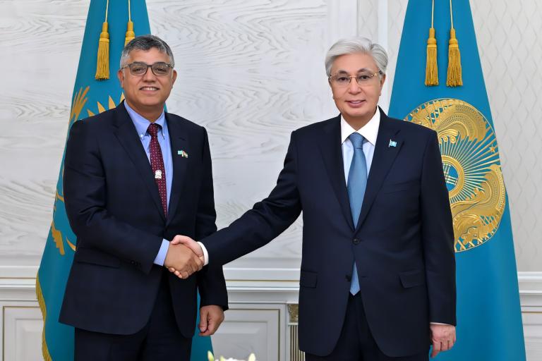 Two men in suits shake hands in front of two blue flags with gold emblems in a formal setting.