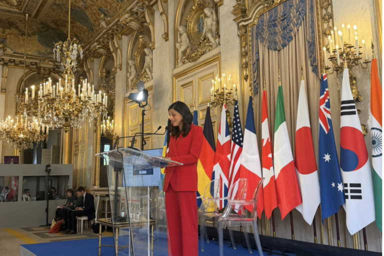 A woman in a red suit speaks at a clear podium in an ornate room with chandeliers and multiple national flags displayed behind her.