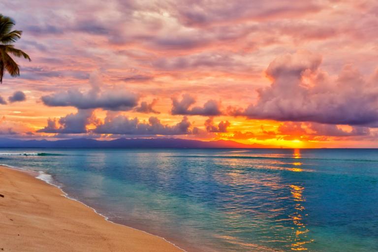 A sandy beach with calm blue water at sunset, palm trees on the left, and colorful clouds in the sky reflecting orange and pink hues.