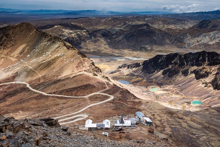 A remote mountainous landscape with winding dirt roads, a cluster of white buildings in the foreground, and rugged, rocky hills and small turquoise lakes in the background.