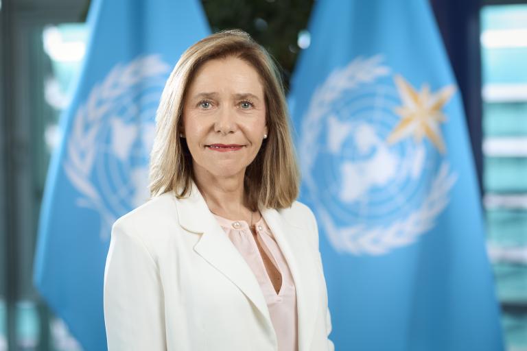 A woman in a white blazer stands in front of two blue flags featuring the United Nations emblem.