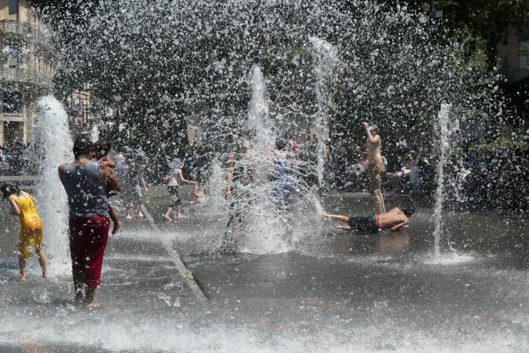 People, including children, play and cool off among multiple water fountains in an urban outdoor setting on a sunny day.