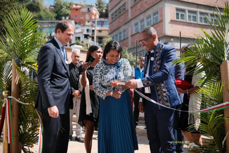 Three people stand smiling at an outdoor ribbon cutting ceremony, surrounded by others and greenery, with buildings visible in the background.