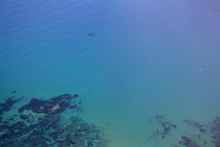 Aerial view of clear blue-green ocean water with dark patches of underwater rocks near the shore.
