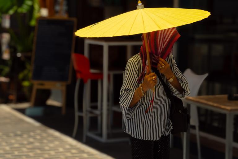 Person holding a large yellow umbrella, face covered by a striped scarf, standing on a sunlit sidewalk near outdoor seating and a blackboard sign.