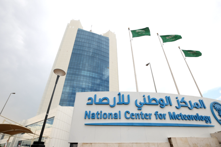A modern glass building stands behind a sign for the National Center for Meteorology, with three Saudi Arabian flags flying nearby.