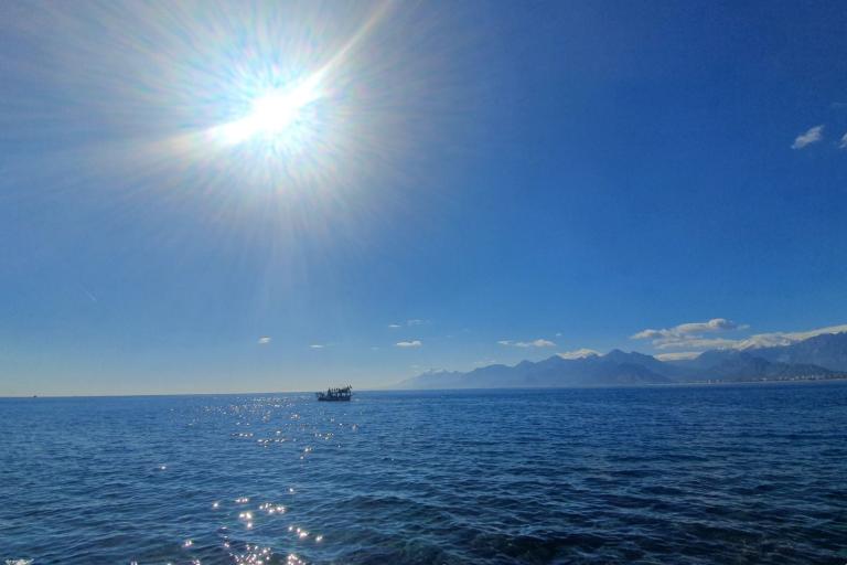 The sun shines brightly over a calm blue sea with a boat in the distance and mountains visible on the horizon under a clear sky.