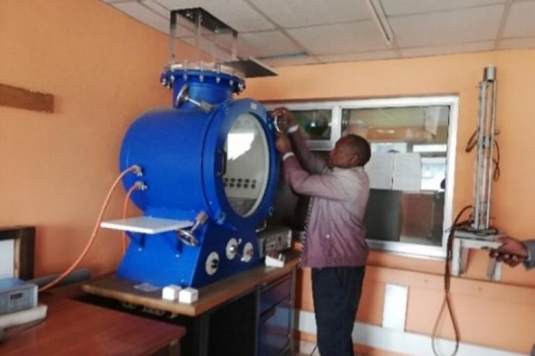 A person adjusts controls on a large blue scientific instrument in a laboratory with light orange walls and various equipment.