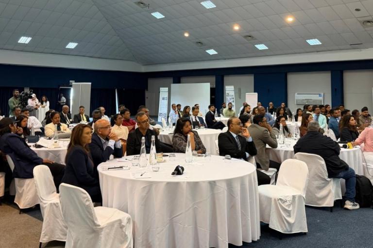 People seated at round tables in a large conference room, attentively listening to a presentation. The room is well-lit with white tablecloths and covered chairs.