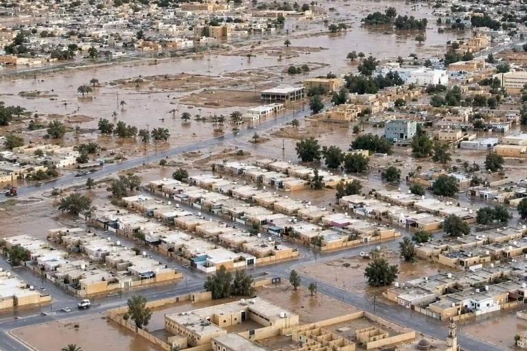 Aerial view of a town affected by flooding, with water covering streets and partially submerging buildings; desert landscape in the background.