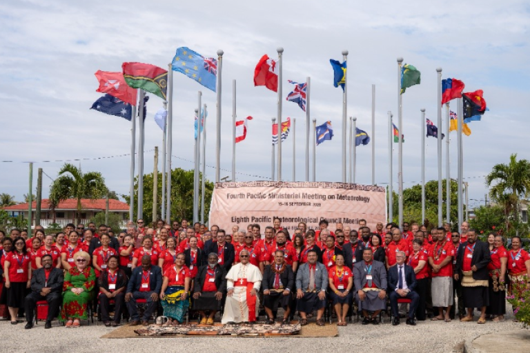 A large group of people pose for a photo in front of international flags and a banner for the Pacific Ministerial Meeting on Meteorology.