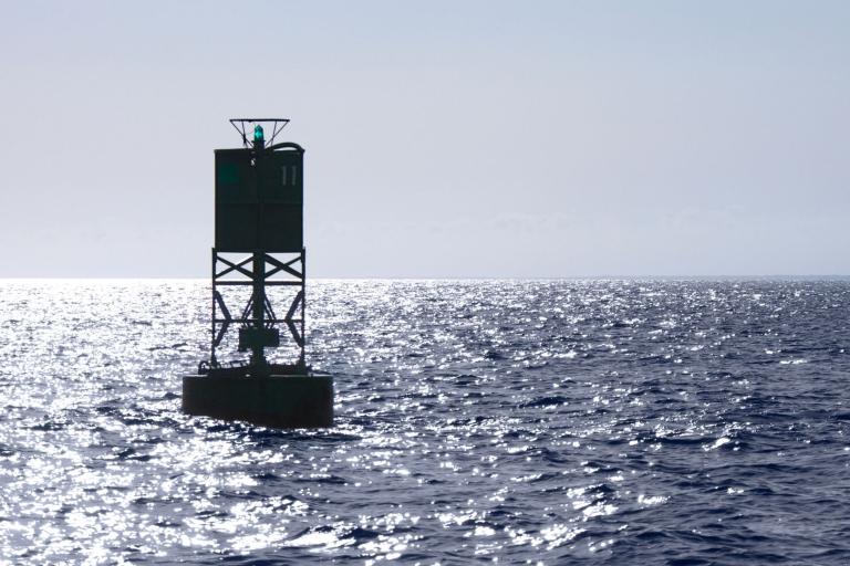 A green navigational buoy with a small light is floating in the open sea under bright sunlight and a clear sky.