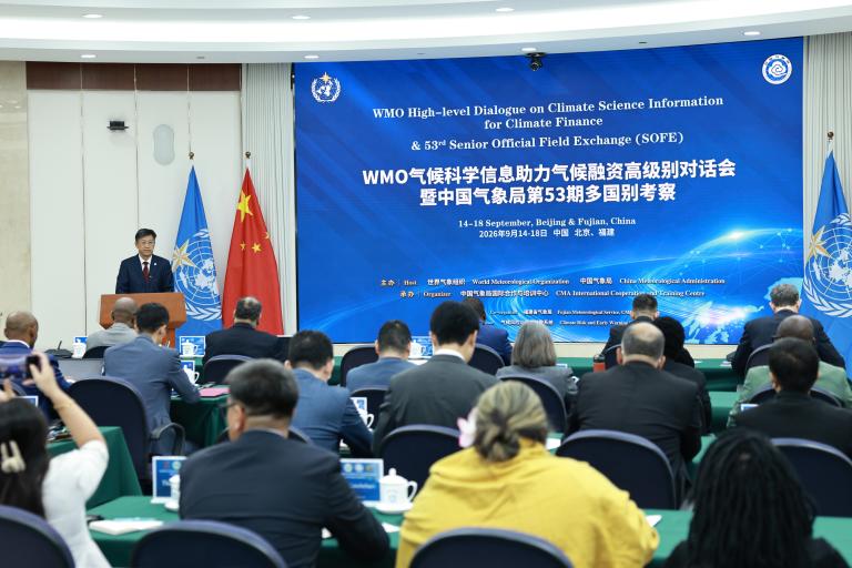 A conference room with delegates seated, facing speakers at a podium under banners for a WMO climate event in Beijing. Chinese and UN flags are displayed behind the speakers.