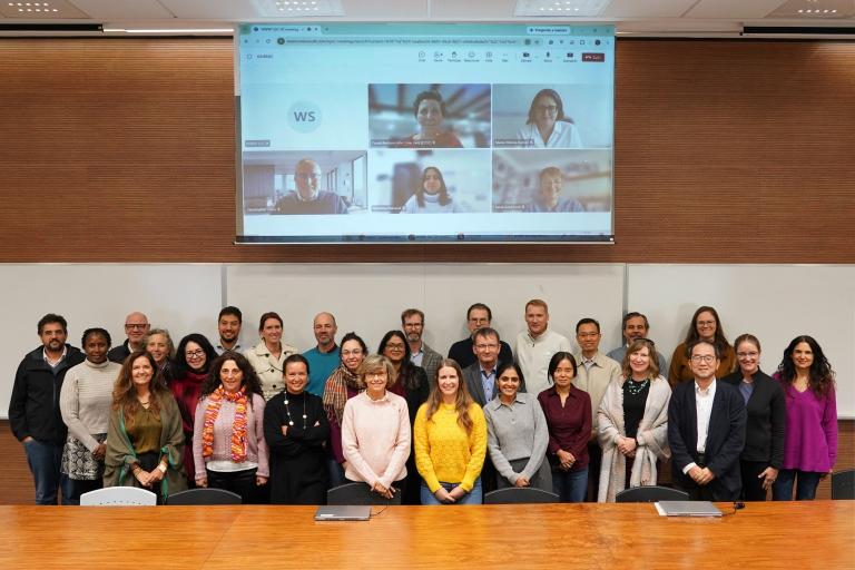 A group of people poses for a photo in a conference room, with a screen behind them displaying a video call with six participants.