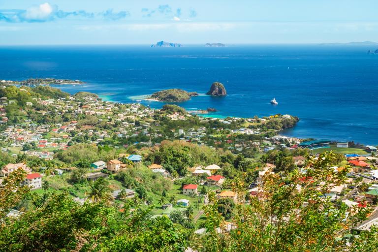 A high-angle view of a coastal town on a hillside overlooking a bay with islands and a yacht in the distance.