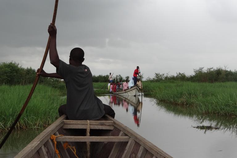 A person steers a wooden boat with a long pole through a grassy channel, following another boat filled with people.
