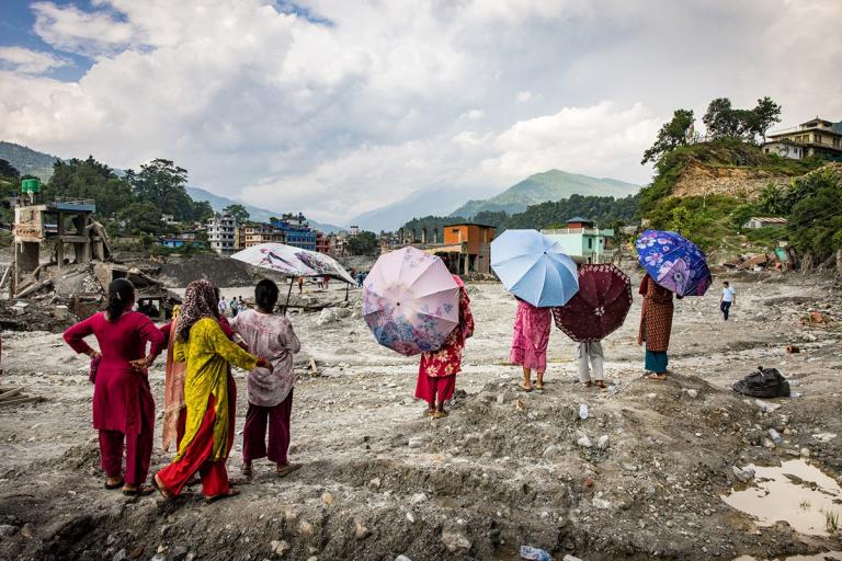 People holding umbrellas stand on a rocky, debris-filled landscape overlooking a town near mountains.