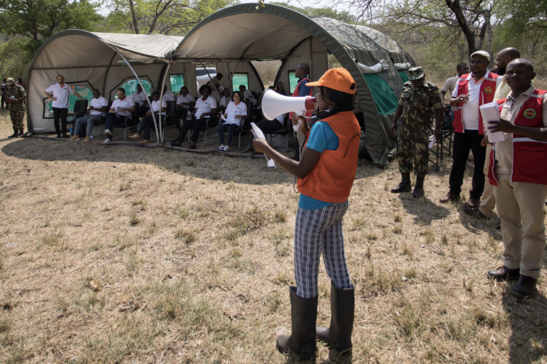 A person in an orange vest speaks through a megaphone to a group of people seated under a large canvas tent in a field.