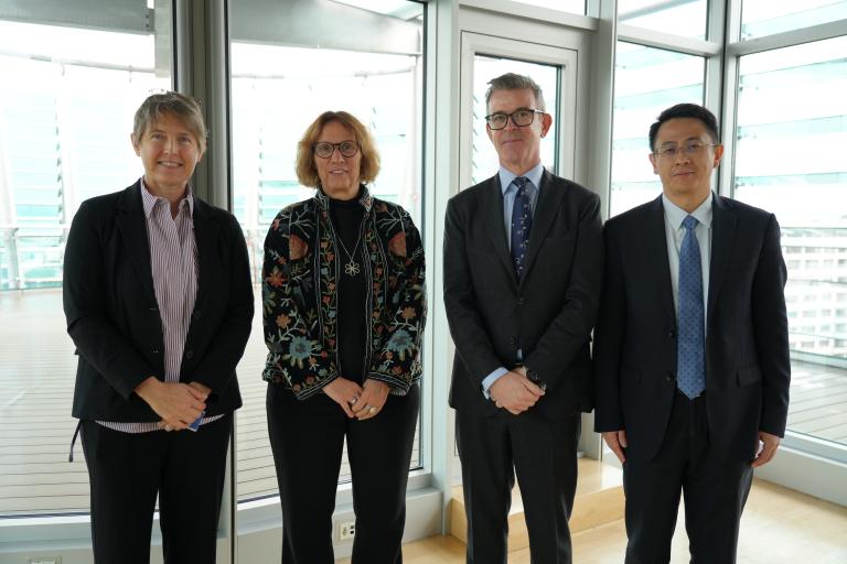 Four people in business attire standing in a row in a bright, modern room with large windows.