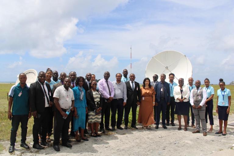 A group of people poses outdoors in front of satellite dishes on a grassy area, dressed in business and casual attire under a partly cloudy sky.