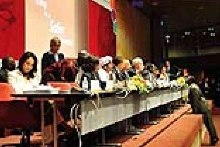A group of people sits at a long table on a stage during a formal conference or panel discussion, with microphones and documents in front of them.