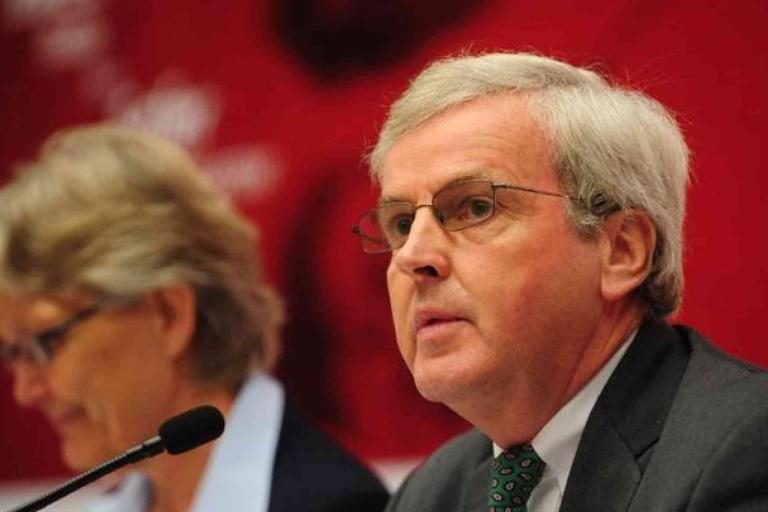 Two people sit at a table with microphones; the man in the foreground wears glasses and a suit, and the background is red and slightly blurred.