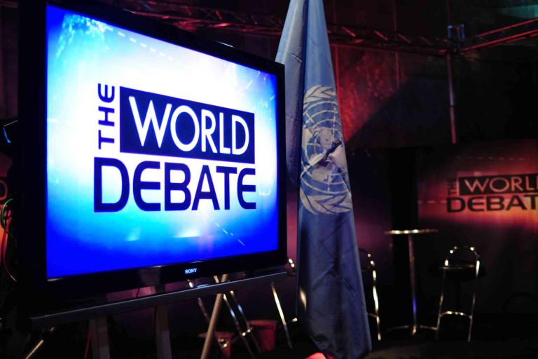 A television screen displays “The World Debate” with a United Nations flag and chairs visible in a dimly lit studio setting.